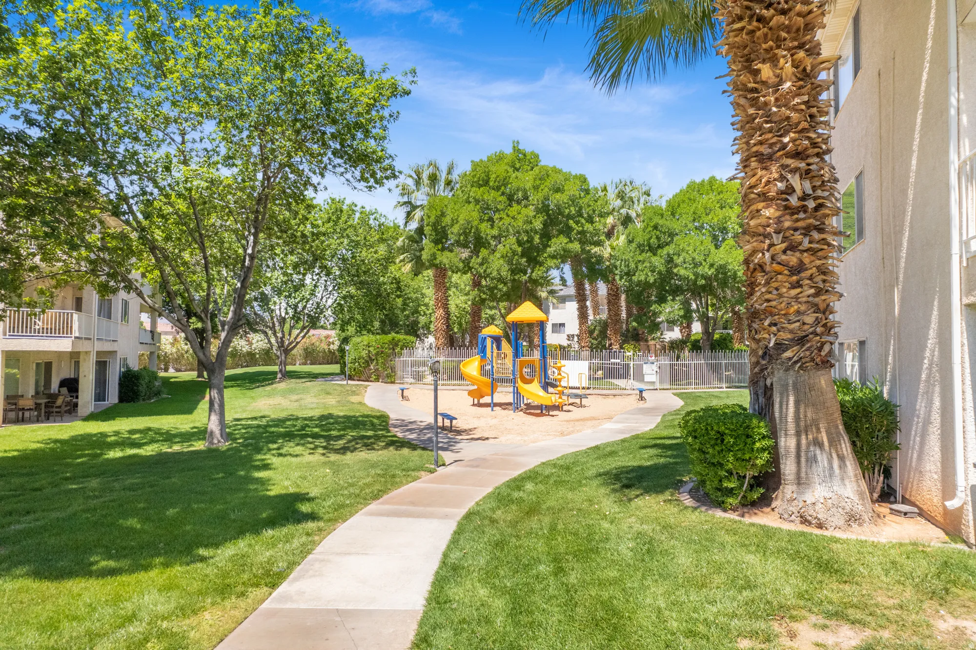 Landscaped outdoor area featuring a playground with slides and climbing structures, a concrete walkway, mature trees, green lawn, and a building exterior with balconies