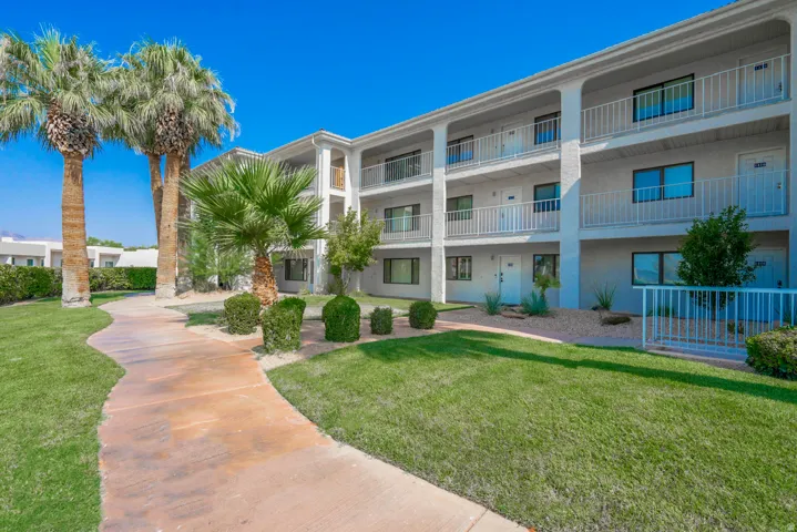 Three-story multi-unit building featuring individual balconies with white railings, a winding concrete pathway, and mature palm trees