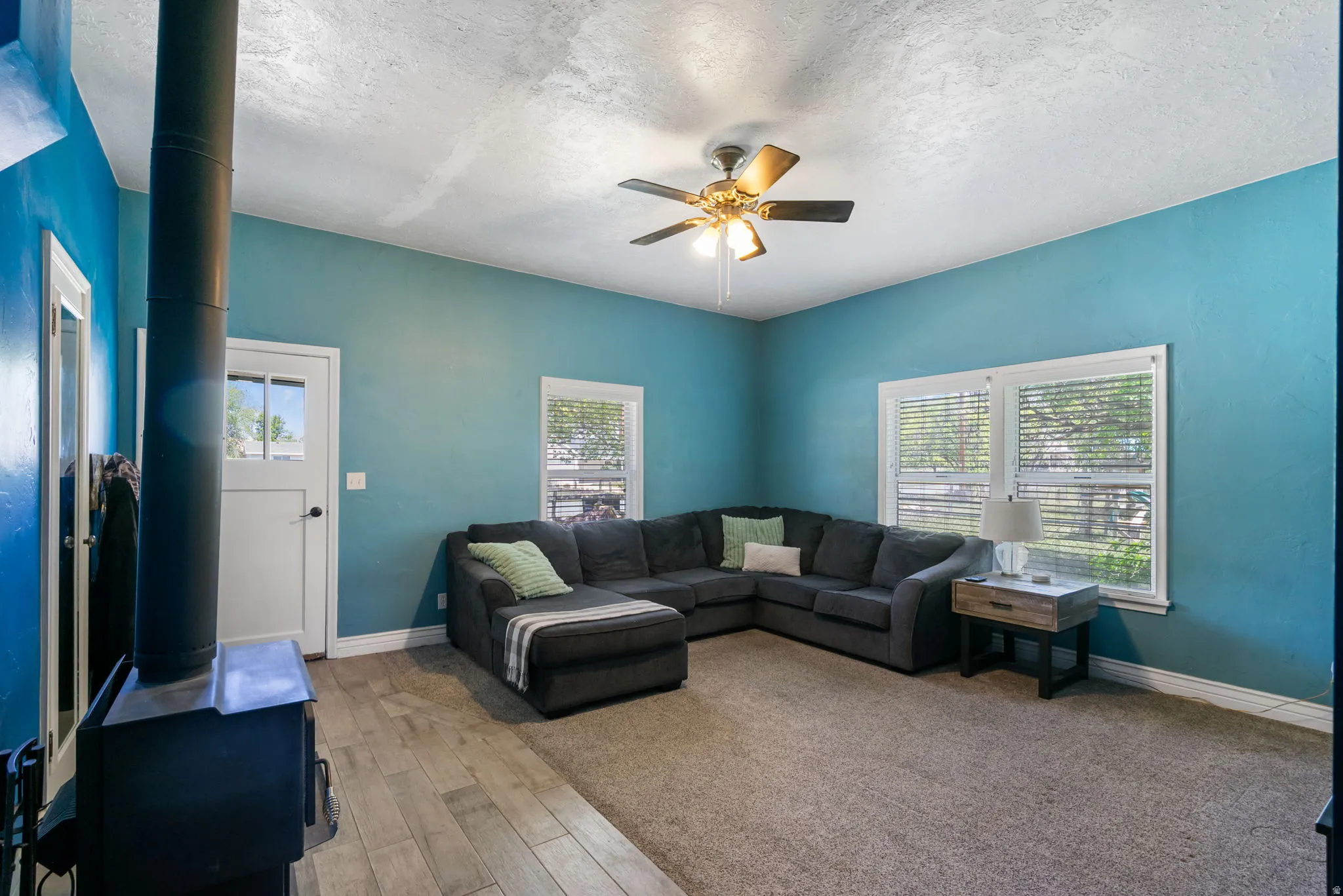 Spacious living area featuring a wood-burning stove, ceiling fan, and two large windows with blinds