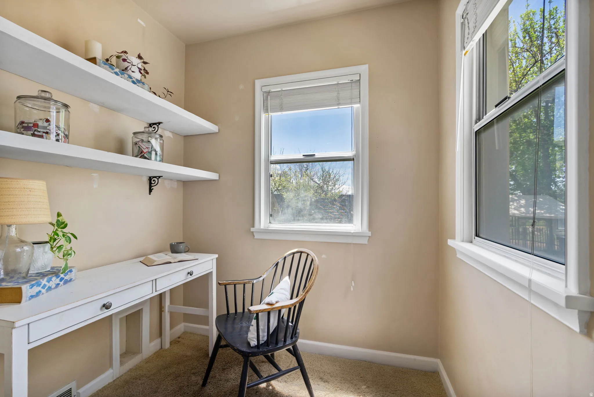 Sunlit room featuring two windows with white trim, built-in white floating shelves with decorative brackets, a built-in white desk with two drawers, and neutral carpeting