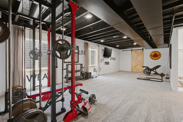 Spacious recreation room featuring light gray carpeting, exposed black ceiling joists and ductwork, a natural wood-finish barn door, and recessed lighting