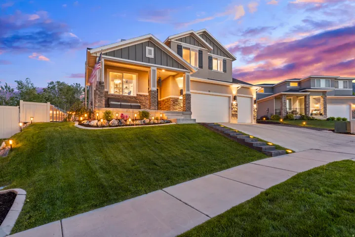 Two-story residence featuring a covered entry porch with stone accents, a dual-bay garage, a manicured lawn, and a paved driveway with integrated lighting