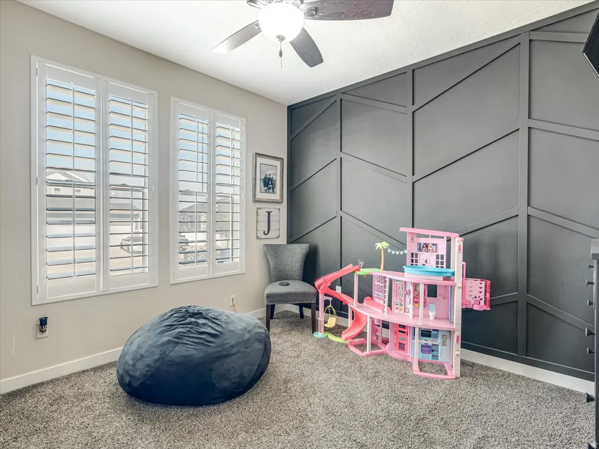 Accent wall with geometric paneling, two windows with white plantation shutters, textured carpeting, and a ceiling fan with integrated lighting