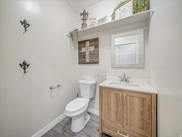 Bathroom featuring a wood-finish vanity with a white countertop and basin sink, a wall-mounted mirror, a toilet, and wood-look flooring
