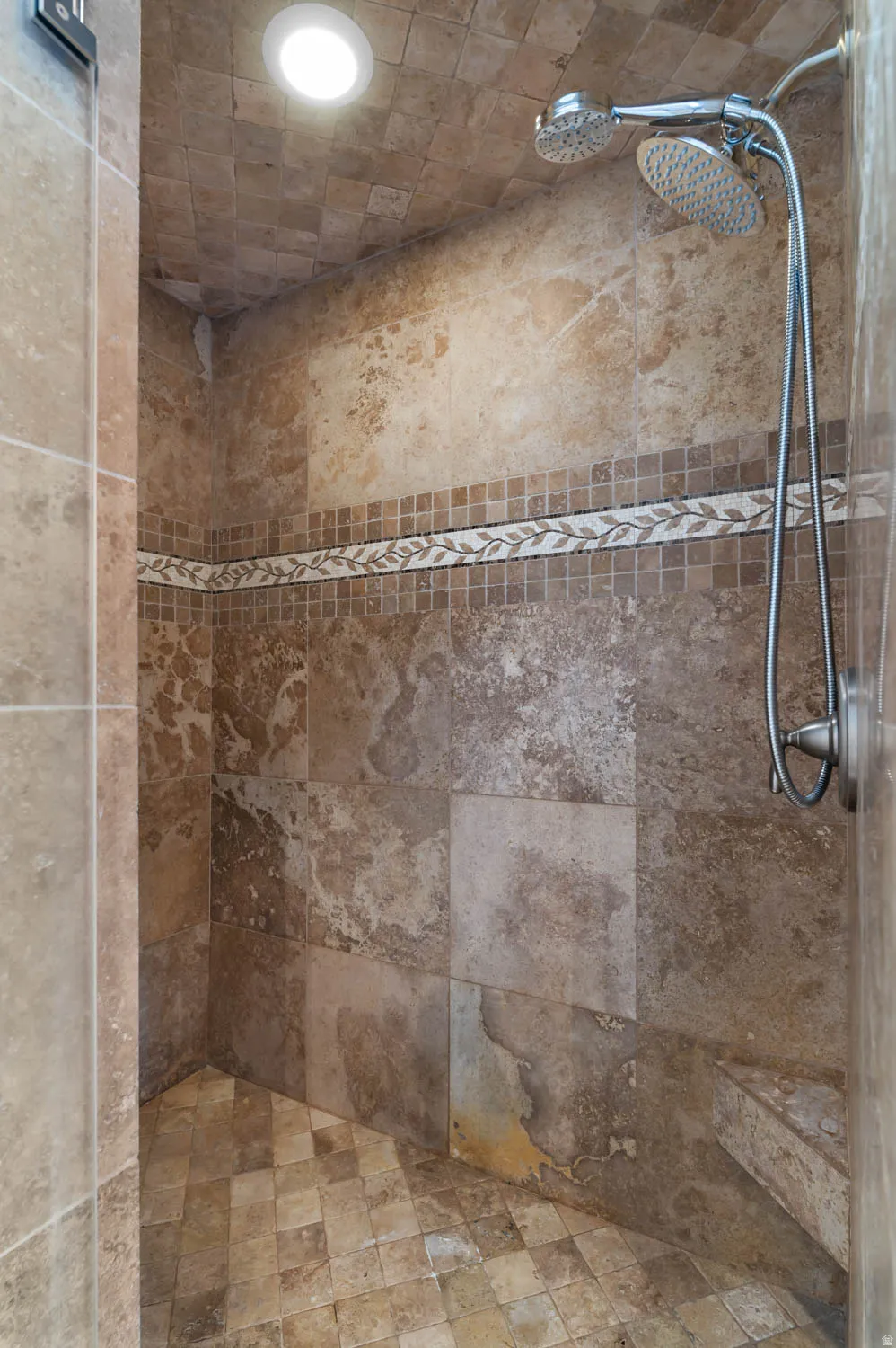 Walk-in shower with natural stone-look tile walls, decorative mosaic accent band, dual shower heads, integrated bench, and recessed lighting