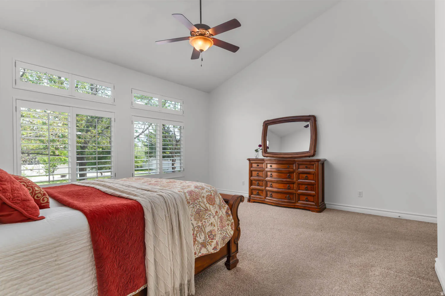 Vaulted ceiling bedroom featuring a ceiling fan with light fixture, carpeted flooring, and plantation shutters on lower windows