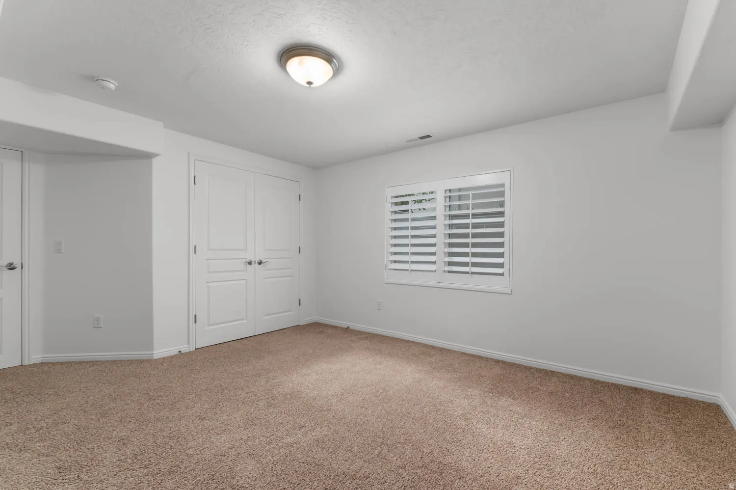 Carpeted interior room featuring a window with plantation shutters, white walls, and bright overhead lighting