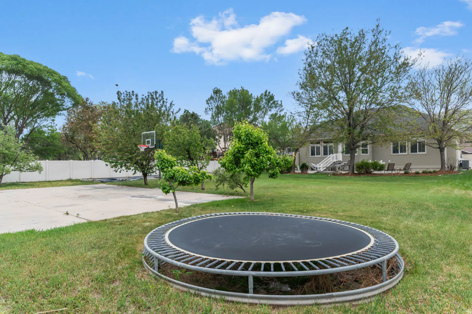 Expansive backyard featuring a lush lawn, mature trees, concrete sports court with basketball hoop, and a vinyl privacy fence