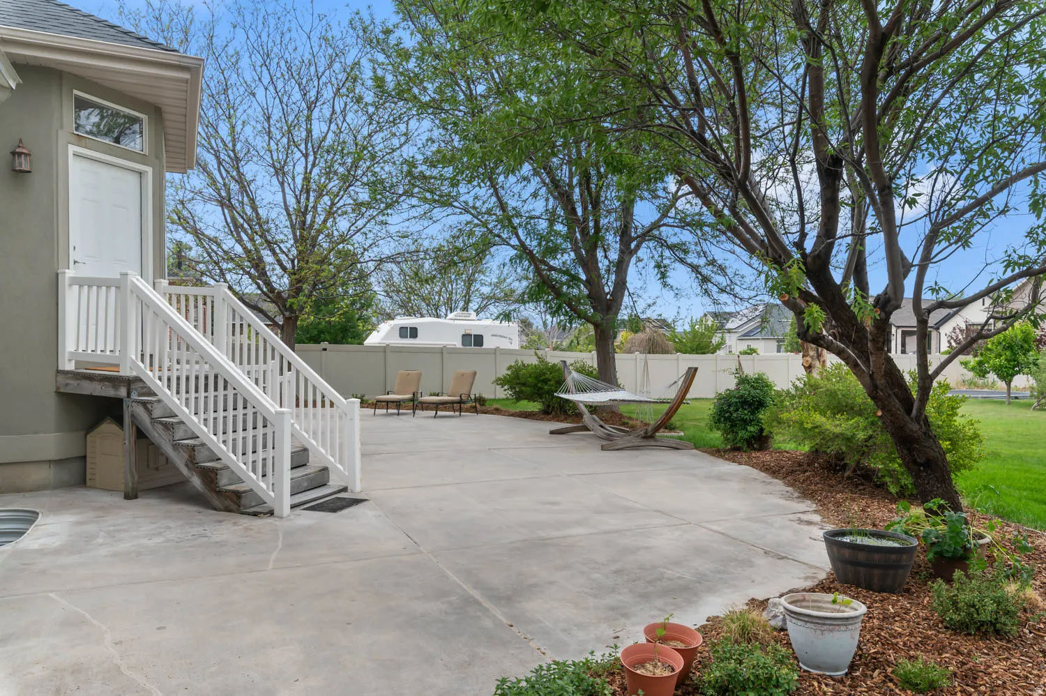 Expansive concrete patio with white railing stairs leading to a side door, framed by mature trees