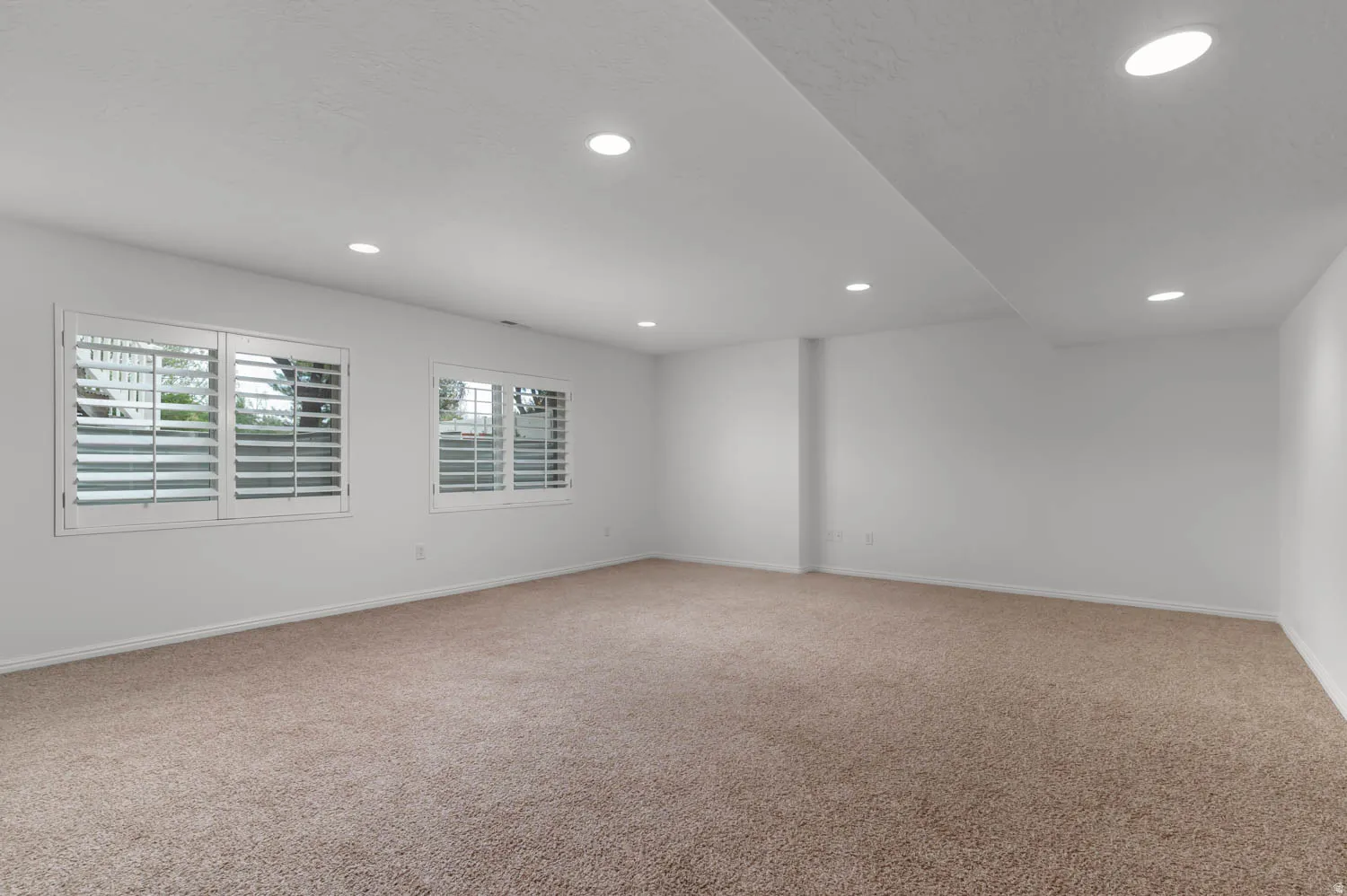 Spacious room featuring neutral carpeting, recessed lighting, and two windows with white louvered shutters