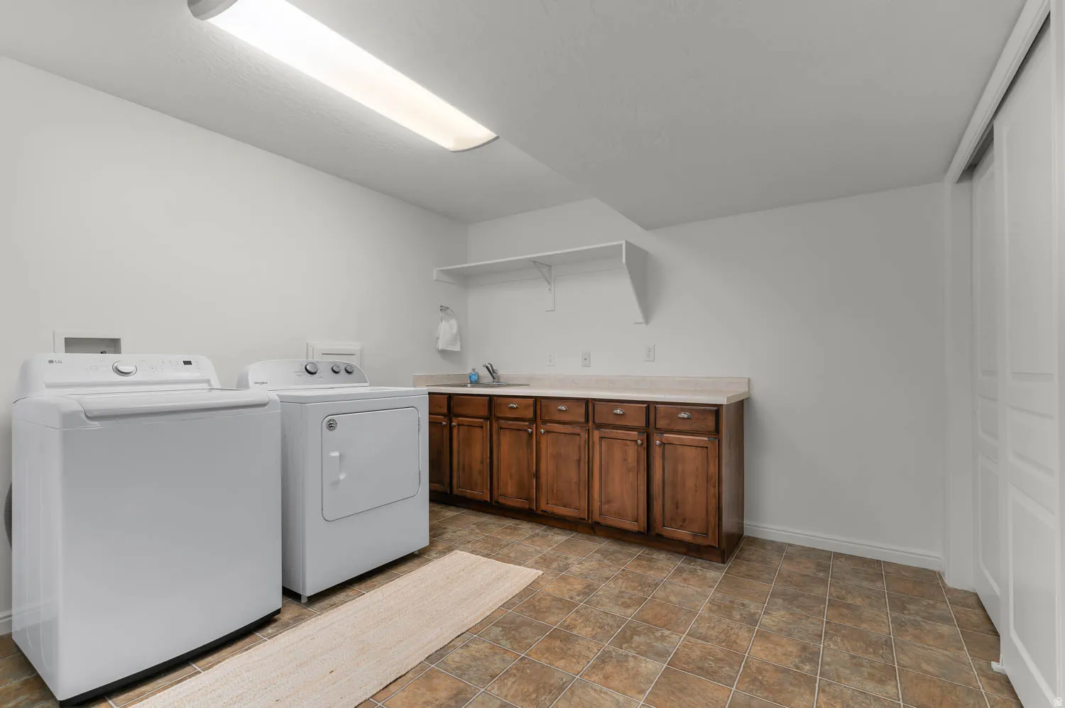 Functional laundry area featuring white walls, tiled flooring, wood-finish cabinetry, a light-toned countertop, and built-in shelving