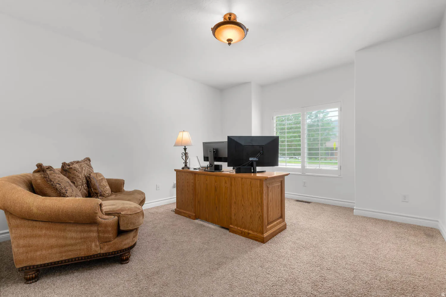 Bright room featuring ample natural light from a window with plantation shutters, neutral-toned carpet, white painted walls, and a decorative ceiling fixture