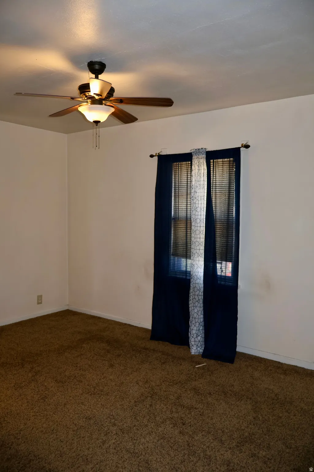 Room featuring wall-to-wall carpeting, white walls, and a window with blinds and dark-colored drapes