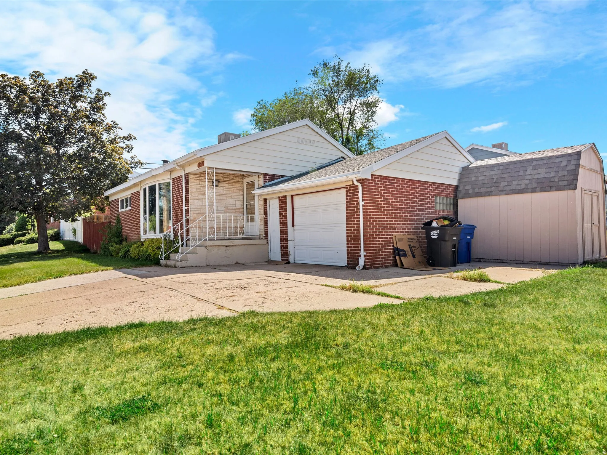 Brick exterior featuring a covered front porch with white railings, an attached single-car garage, an additional detached shed, and a concrete driveway
