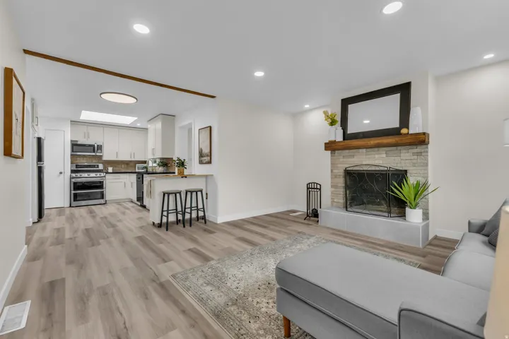 Open-concept living space featuring wood-finish flooring, a stone-surround fireplace with a wood mantle, recessed lighting, and a kitchen area with light cabinetry and a breakfast bar