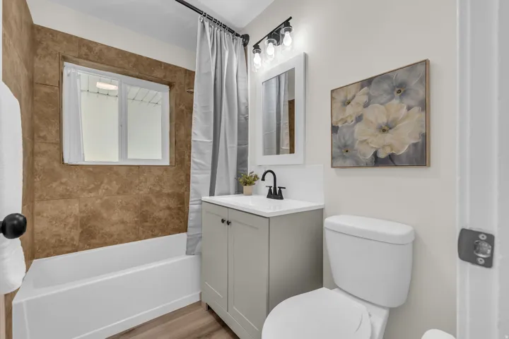 Bathroom featuring a white tub with brown wall tiling, a window, a single vanity with a white countertop and black faucet, and wood-finish flooring