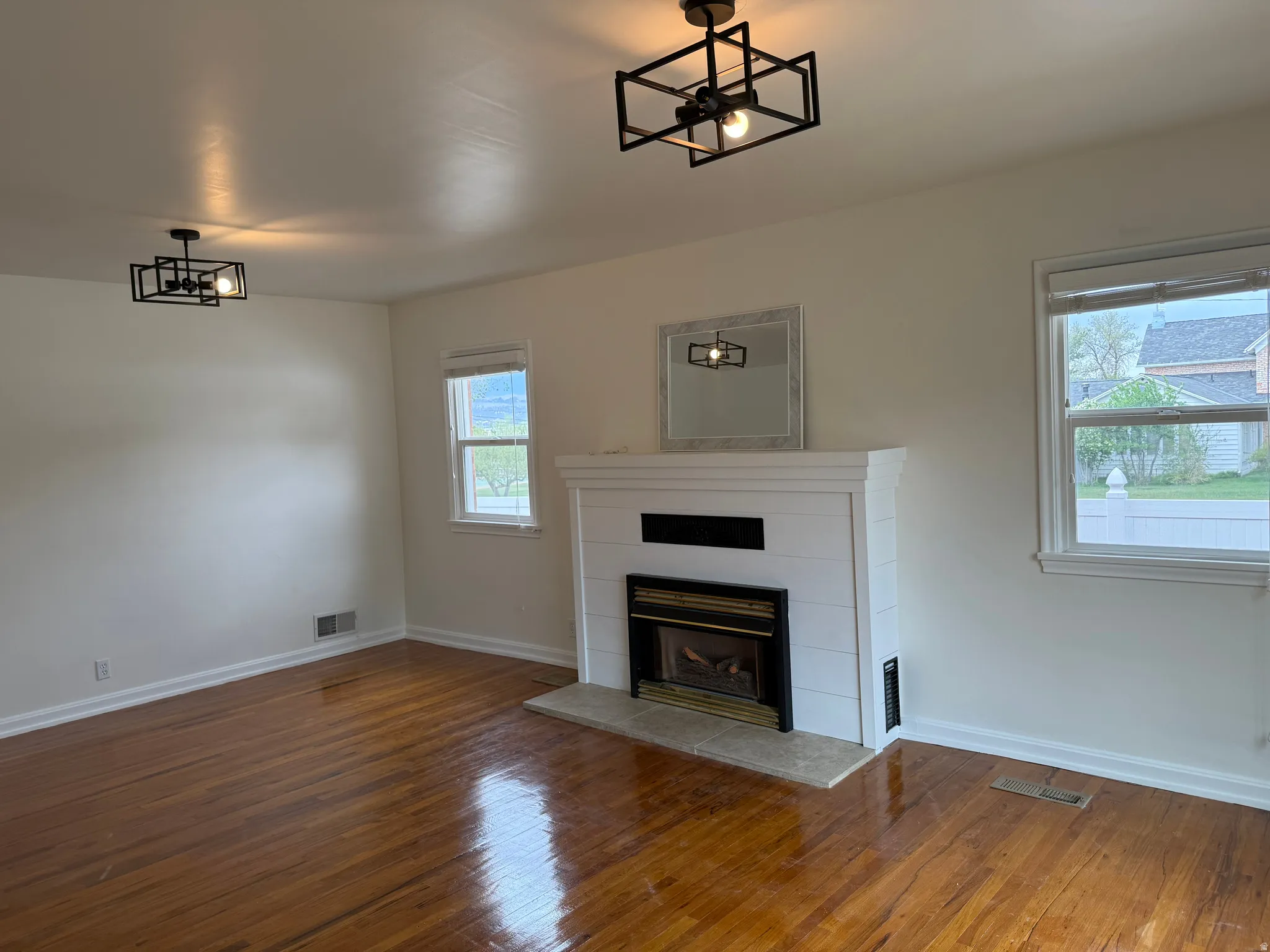 Living area featuring rich wood flooring, a fireplace with a white shiplap-style surround and stone hearth, two windows, and contemporary geometric light fixtures