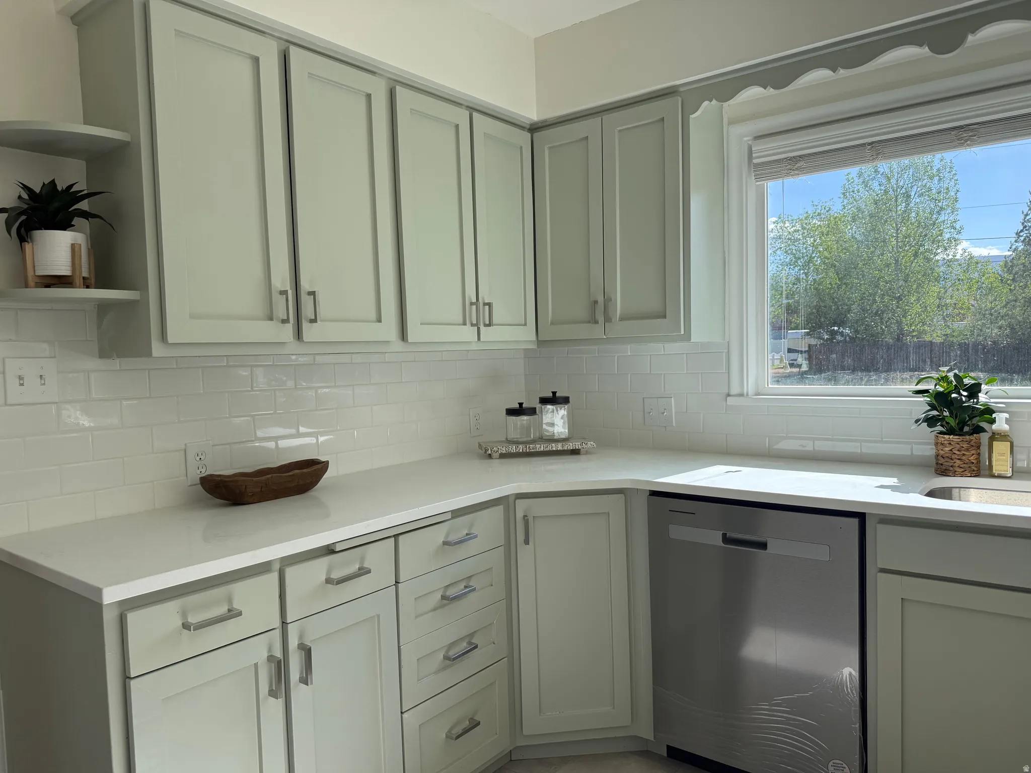 Kitchen featuring light green shaker-style cabinetry, white subway tile backsplash, white countertops, and a stainless steel dishwasher