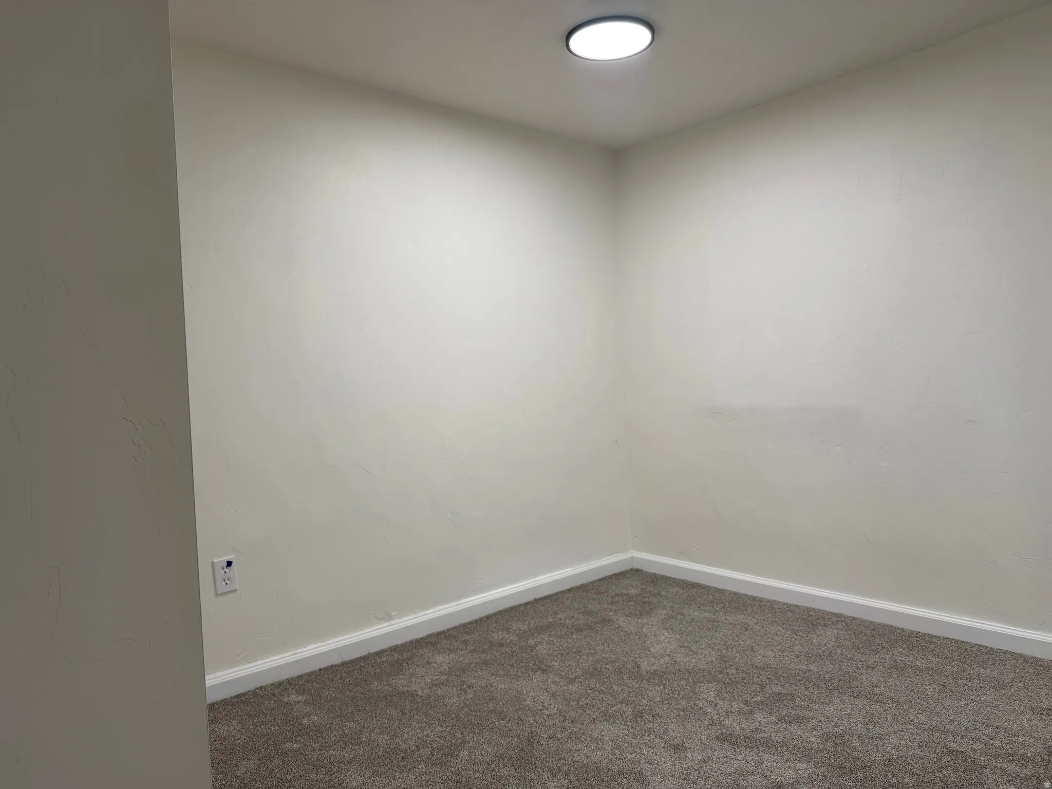 Recessed ceiling lighting illuminates a room with light-colored walls, white baseboards, and gray carpeting