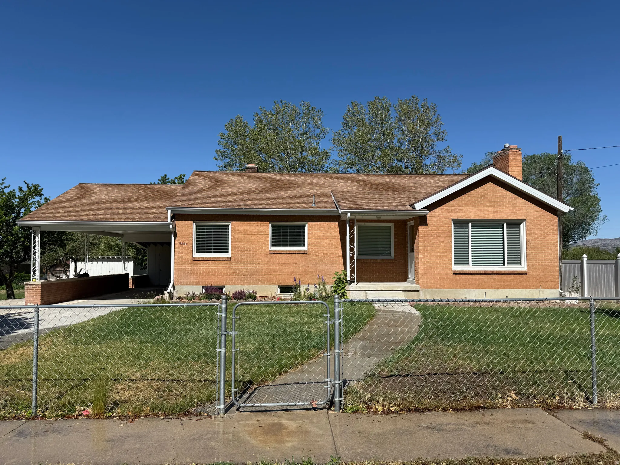 Brick ranch-style home featuring a covered carport, pitched shingle roof, and a front yard with mature landscaping