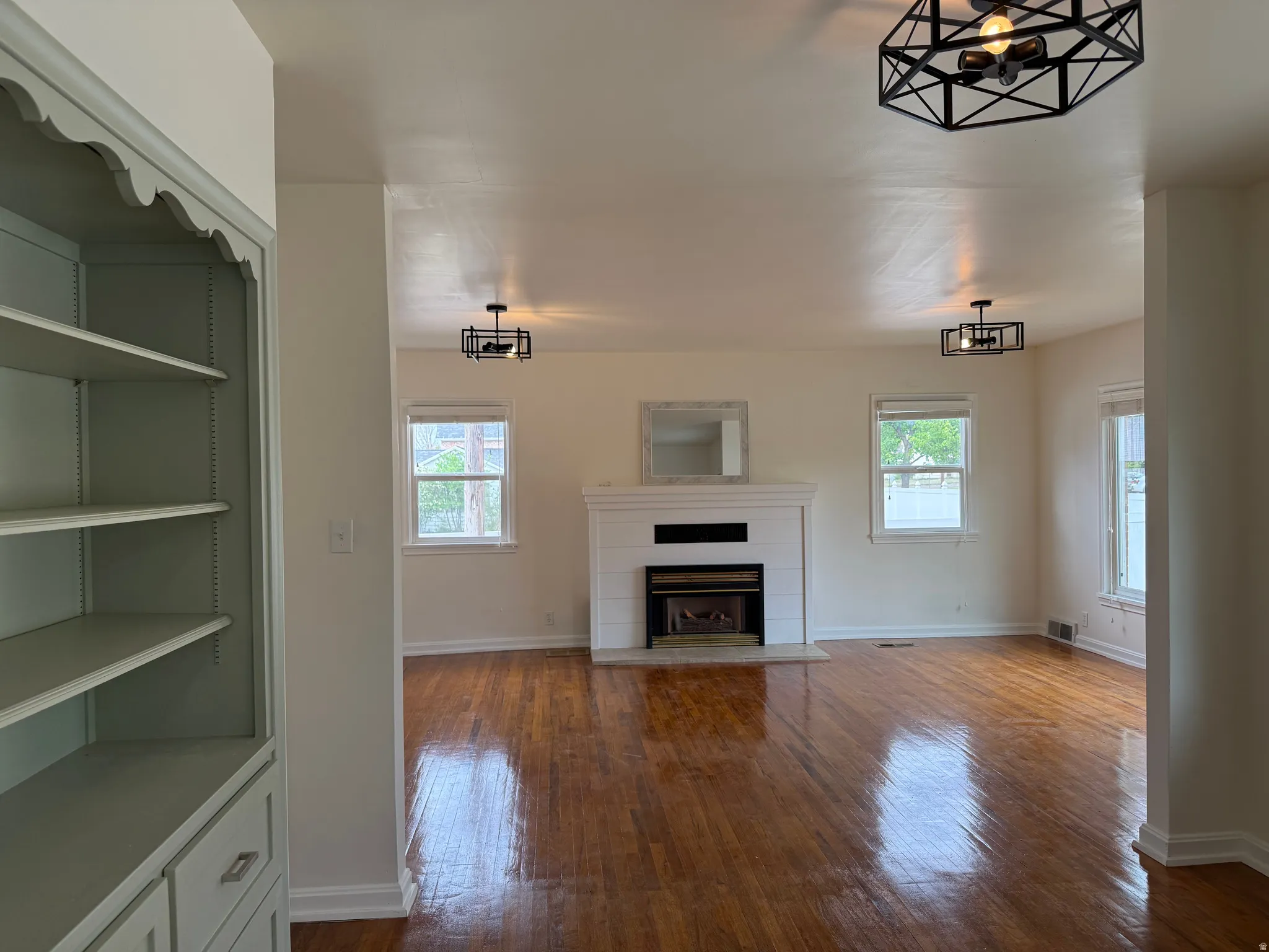 Spacious living area featuring polished wood-finish flooring, a white mantel fireplace, and multiple windows providing natural light
