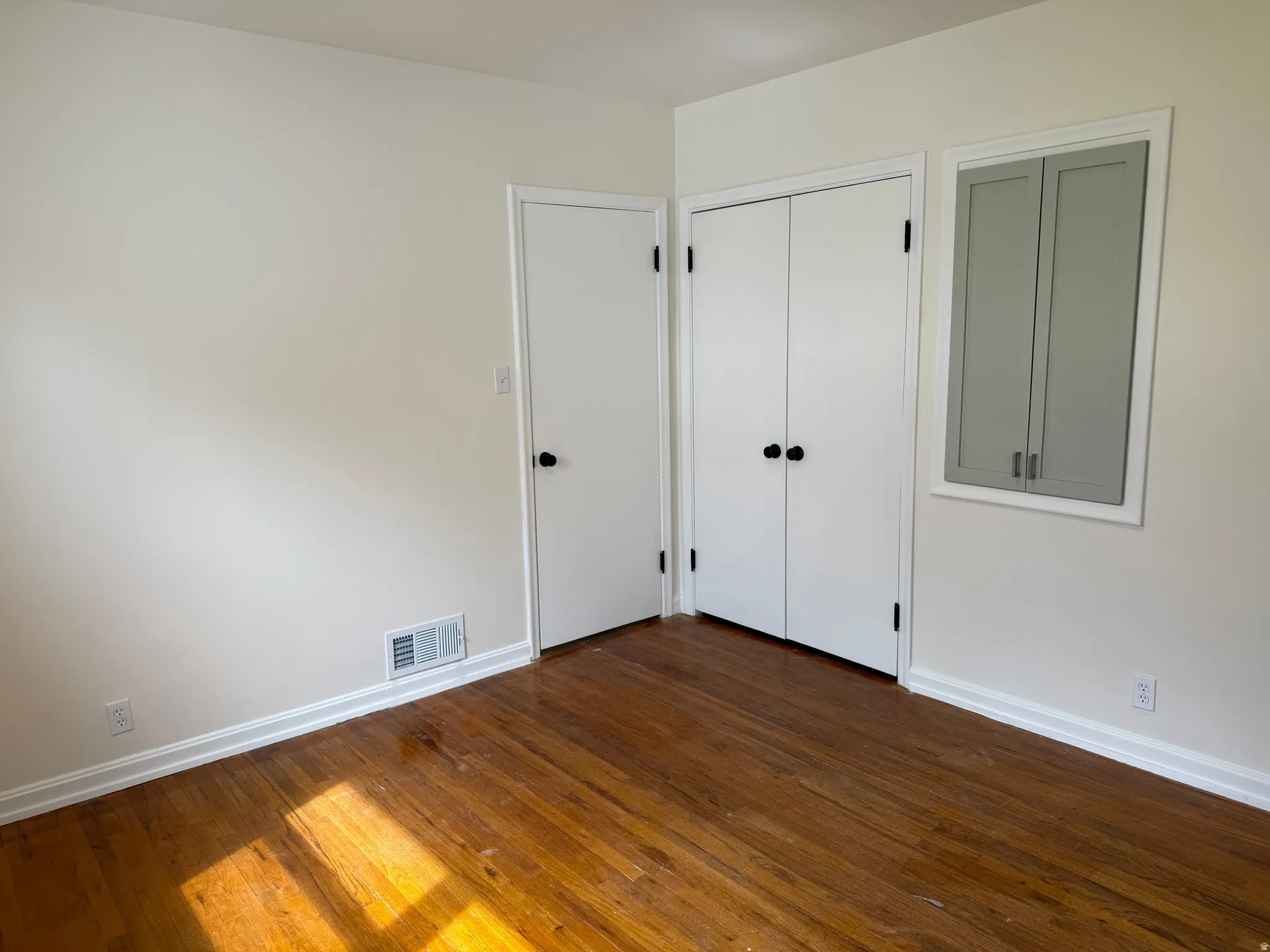 Room featuring wood-finish flooring, white walls, white baseboards, a white door with black hardware, and a built-in cabinetry with gray doors and black hardware
