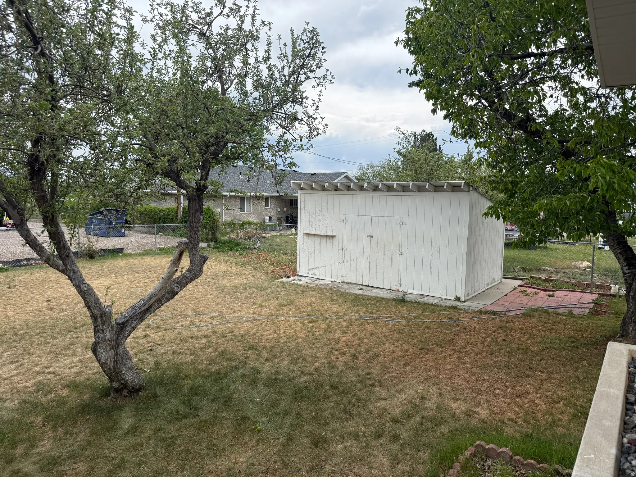 Expansive yard featuring a white storage shed with double doors, established trees, and a concrete pad