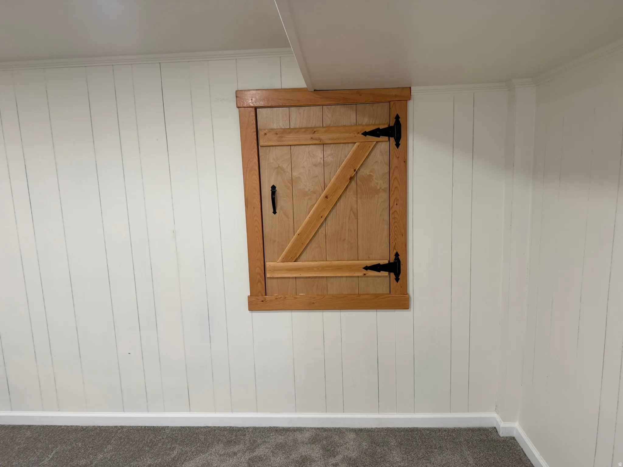Built-in wooden access door with black hardware, complementing paneled walls, white baseboards, and neutral carpeting