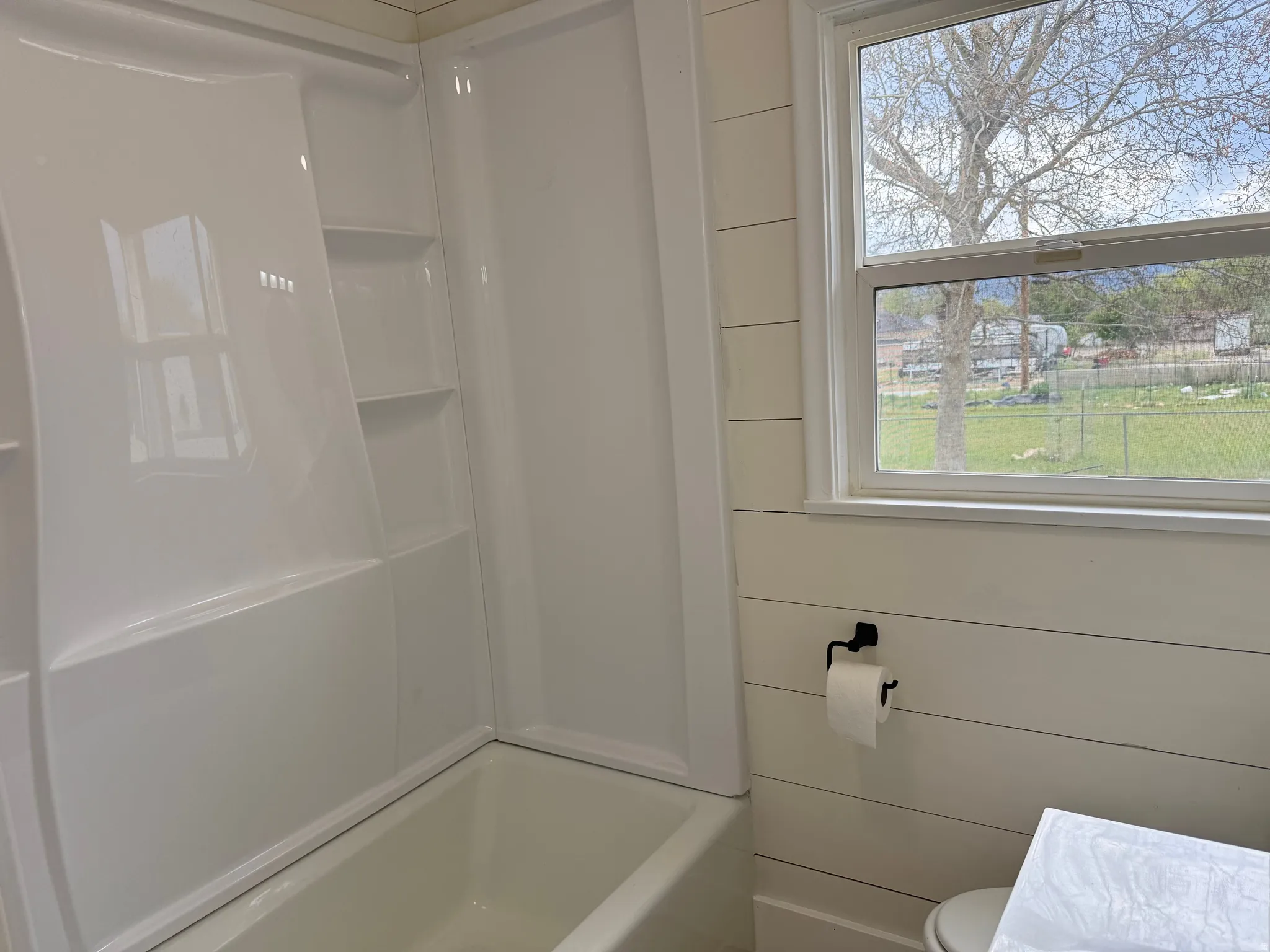 Bright bathroom with a white bathtub and shower surround featuring integrated shelving, a window with exterior views, and shiplap wall paneling
