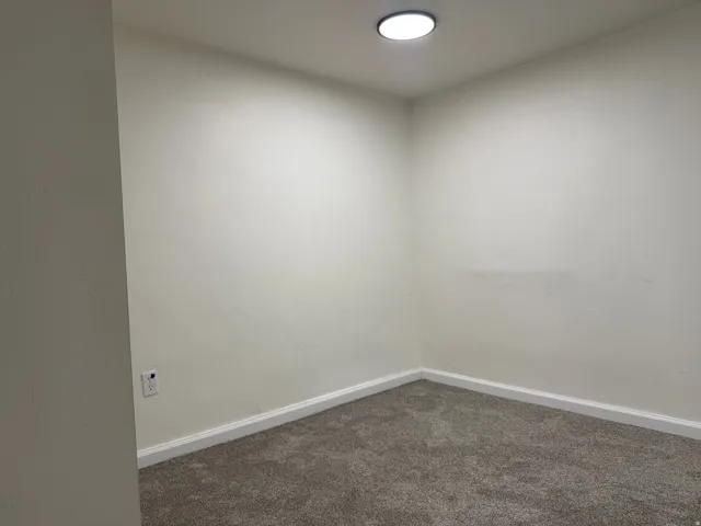 Recessed ceiling lighting illuminates a room with light-colored walls, white baseboards, and gray carpeting