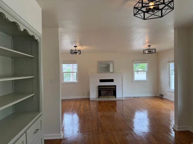 Spacious living area featuring polished wood-finish flooring, a white mantel fireplace, and multiple windows providing natural light