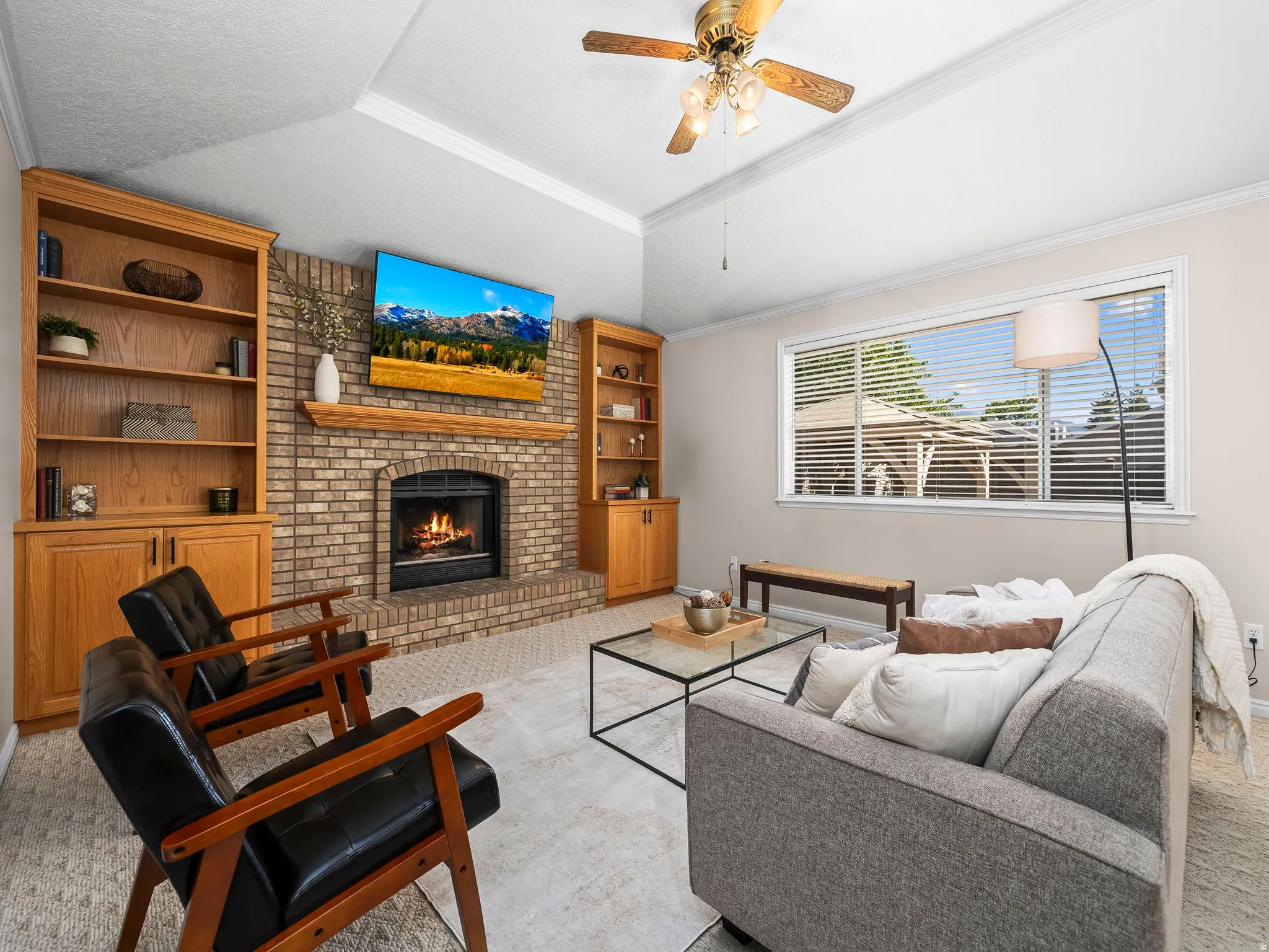 Living area featuring a brick-surround fireplace with a wood mantle, built-in shelving and cabinetry, a ceiling fan, and a large window with horizontal blinds