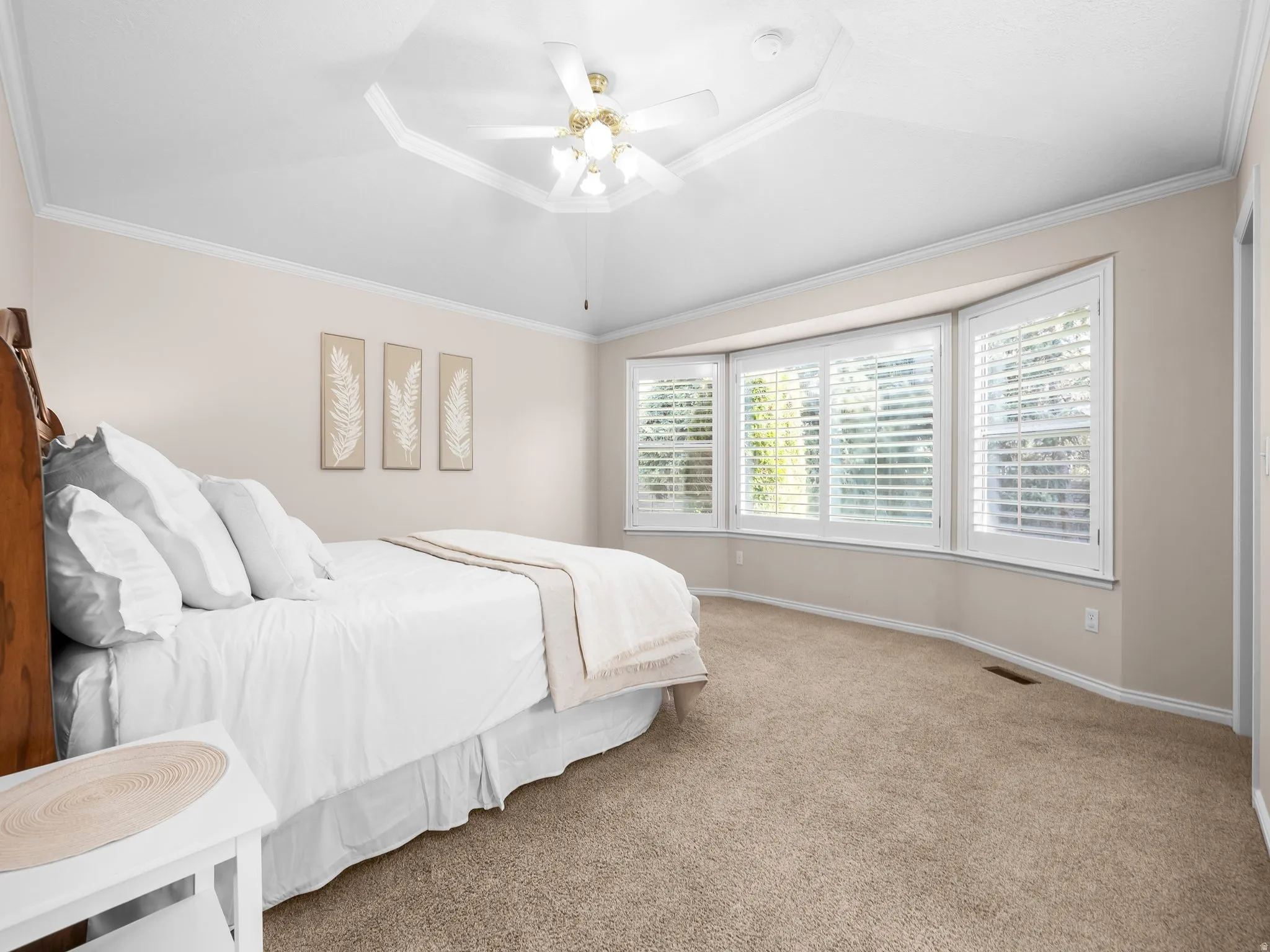 Spacious room with a bay window featuring white plantation shutters, crown molding, a tray ceiling, and a ceiling fan with integrated lighting
