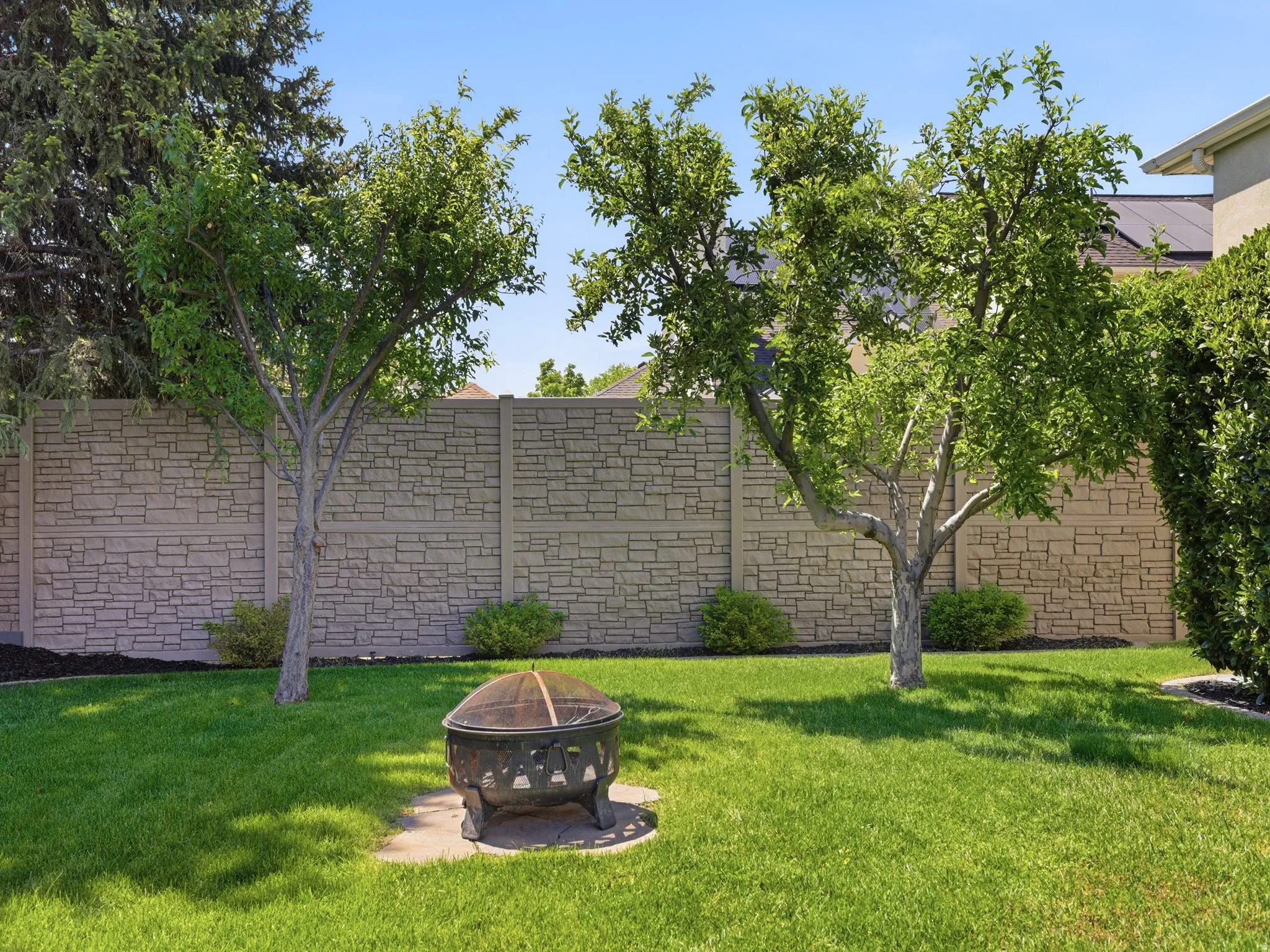 Lush green lawn with mature trees and a decorative fire pit on a stone base, bordered by a textured stone-patterned privacy fence