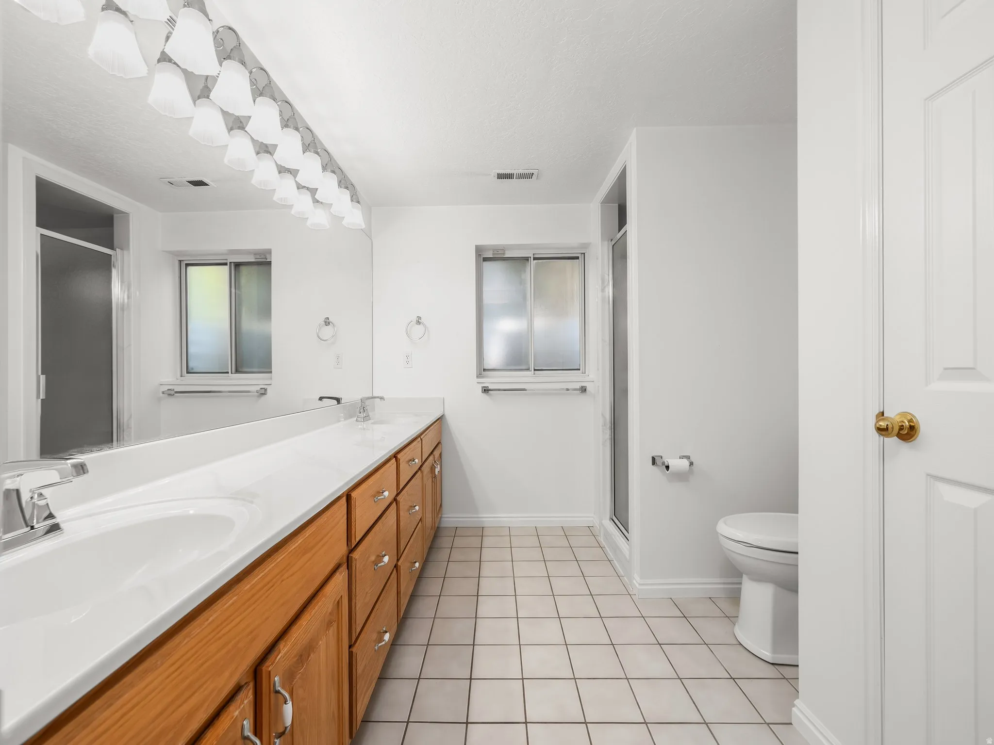 Spacious bathroom featuring a dual vanity with wood-finish cabinetry and a single-piece countertop, a large wall-mounted mirror, and white square floor tiles