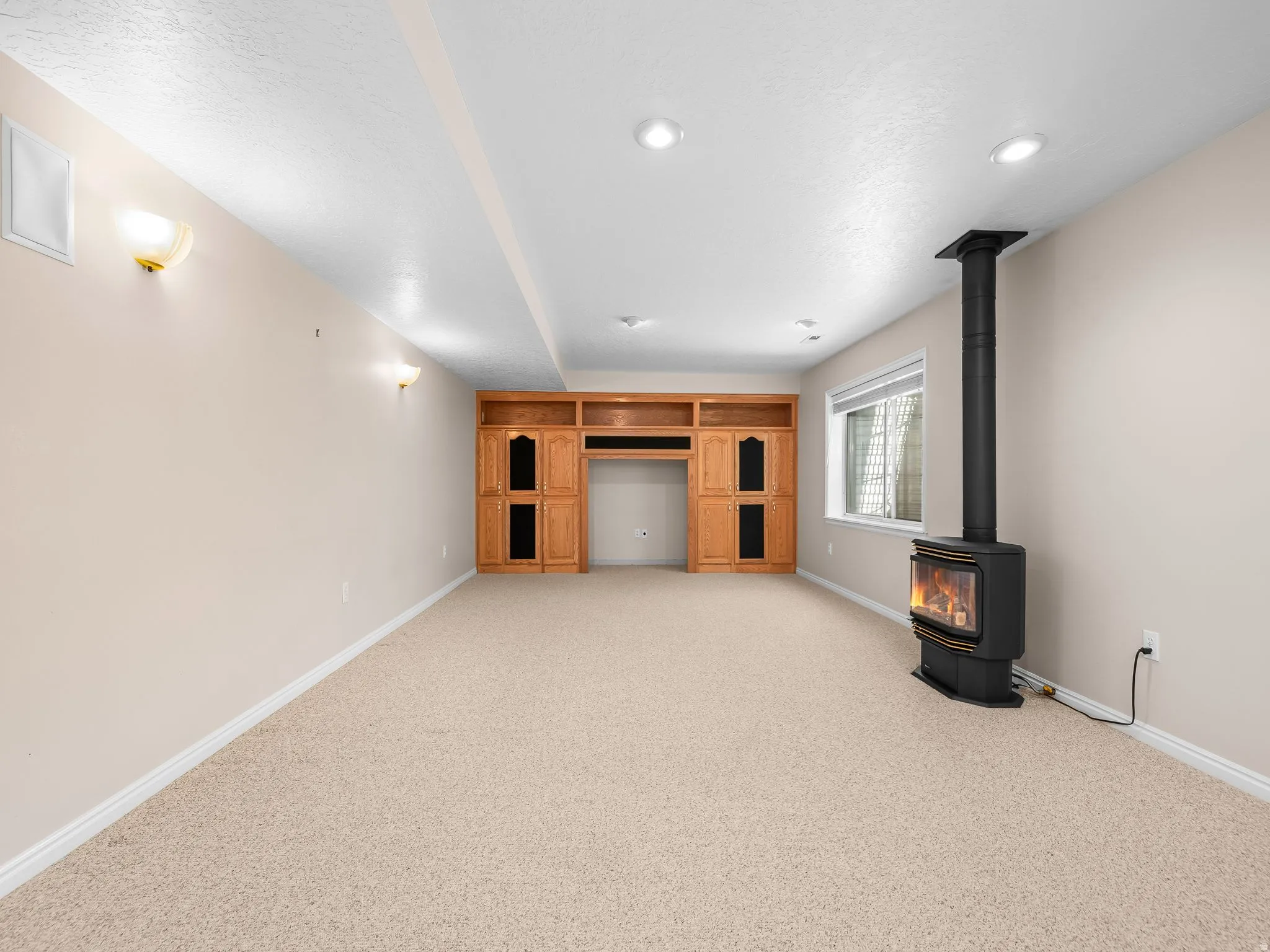Spacious room featuring light beige carpeting, recessed ceiling lighting, and a prominent freestanding black stove with a visible flame