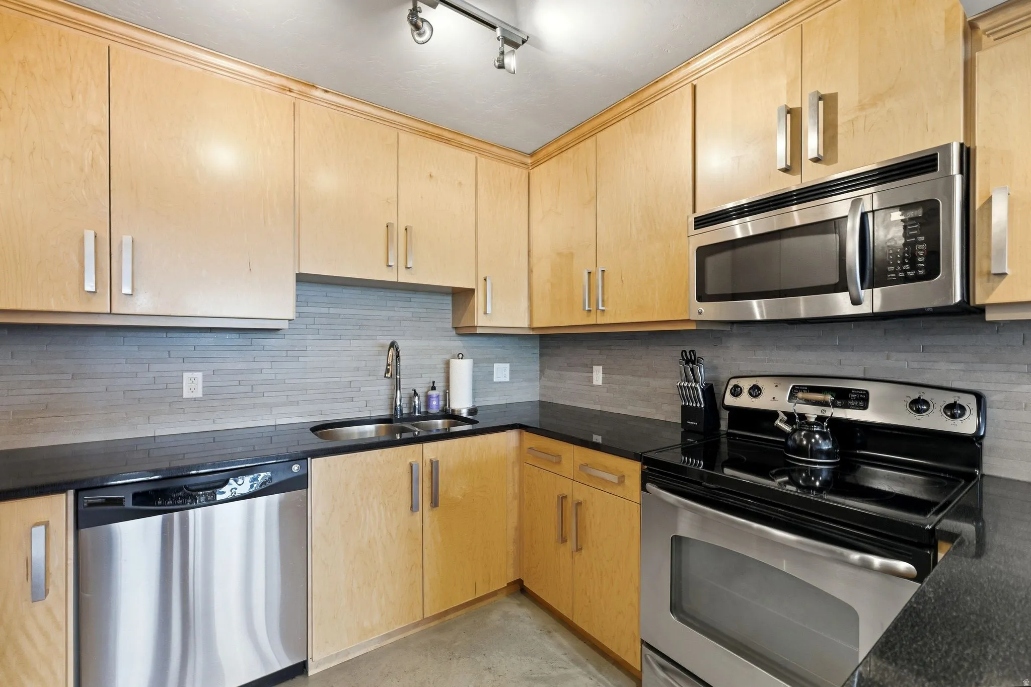 Kitchen featuring light wood cabinetry, dark countertops, a stainless steel sink, a stainless steel dishwasher, and a stainless steel oven with an above-range microwave