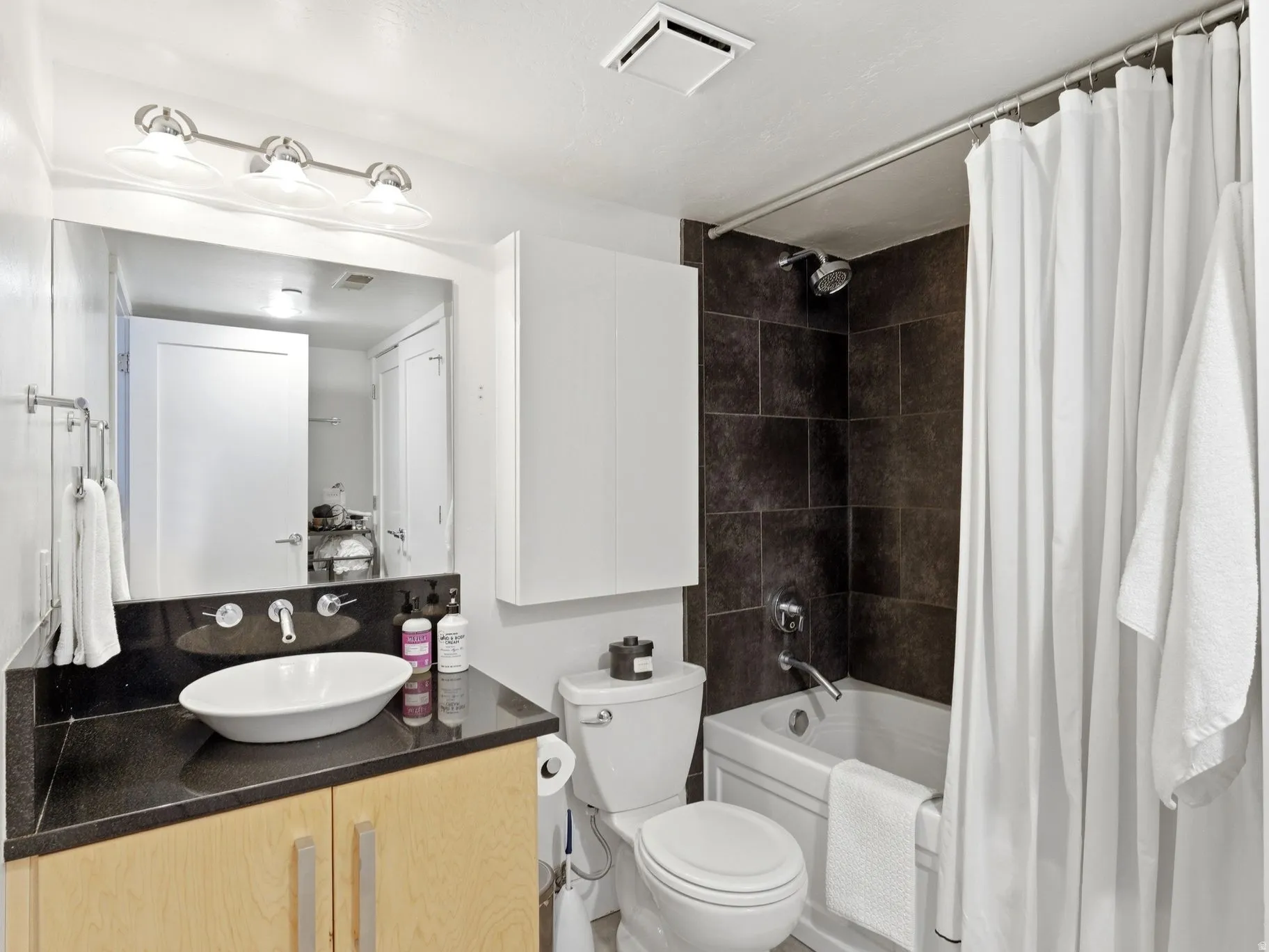 Bathroom featuring a vessel sink atop a dark countertop, light wood-finish cabinetry, a built-in tub with dark tile surround, a wall-mounted faucet, and overhead vanity lighting