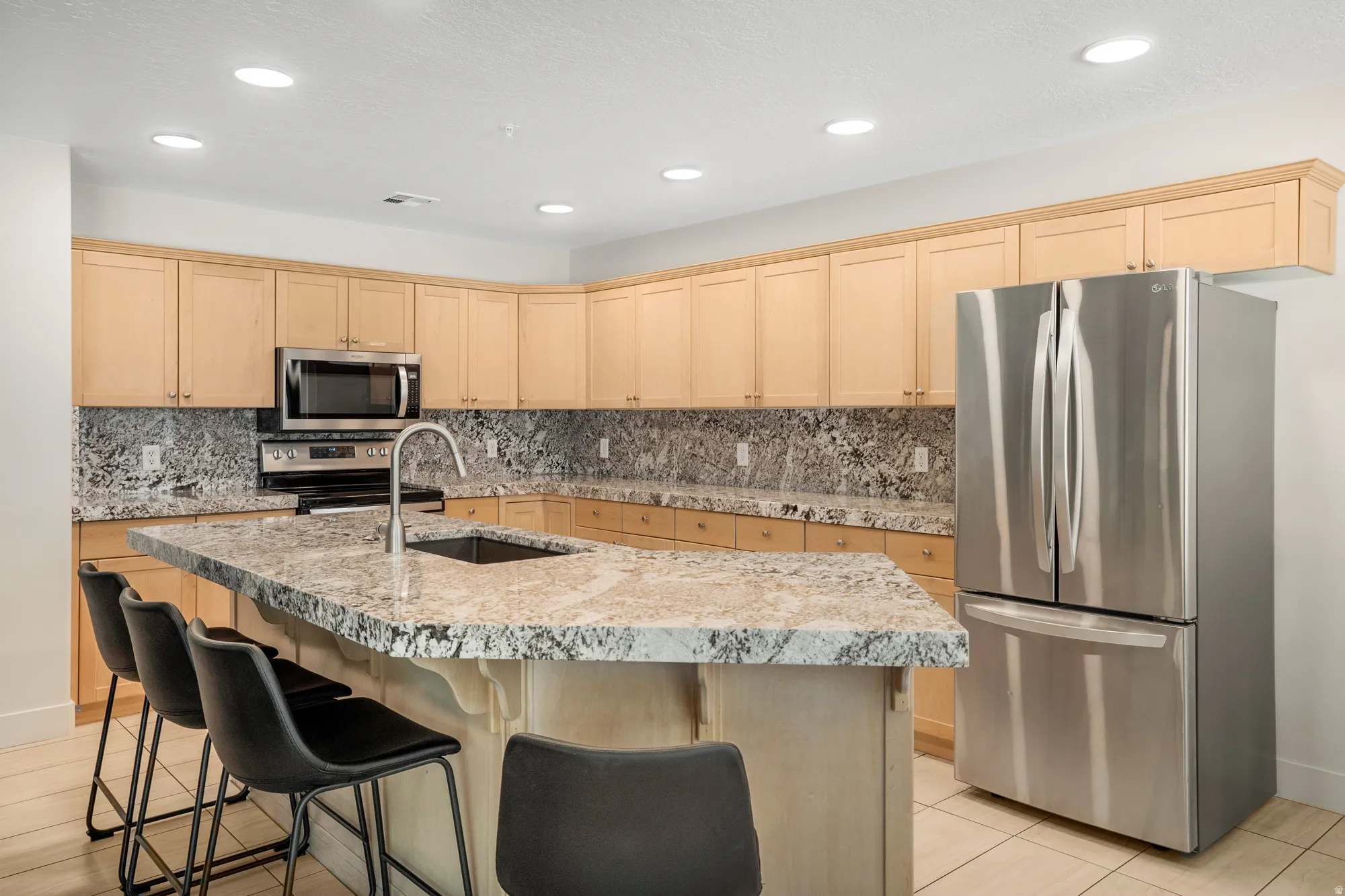 Kitchen featuring a large central island with seating, stone countertops, a stainless steel sink with a gooseneck faucet, light wood cabinetry, and stainless steel appliances including a refrigerator, microwave, and range