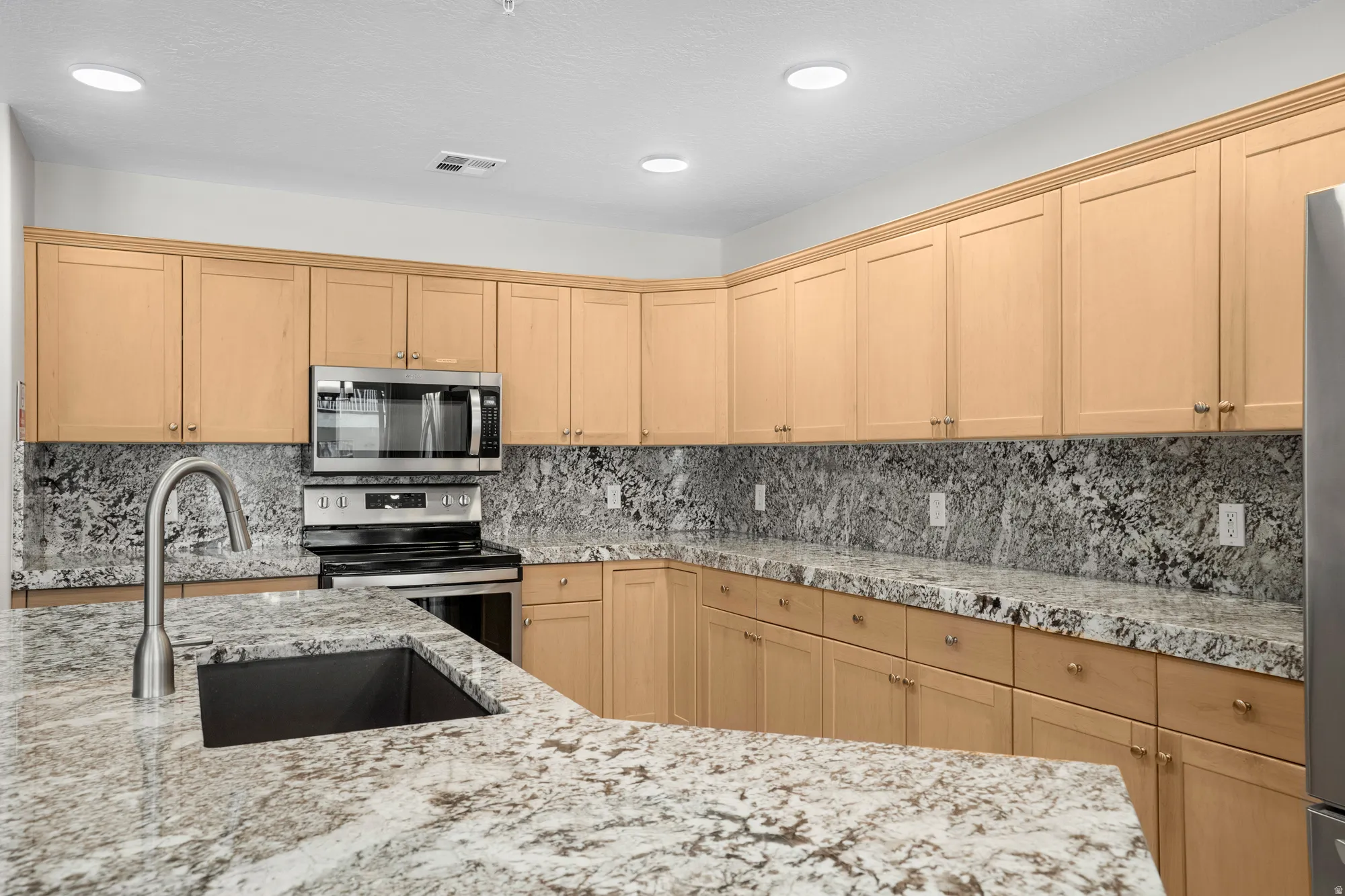 Kitchen featuring a stainless steel undermount sink with a gooseneck faucet, light wood cabinetry, granite countertops, a matching granite backsplash, and stainless steel appliances including a microwave and range