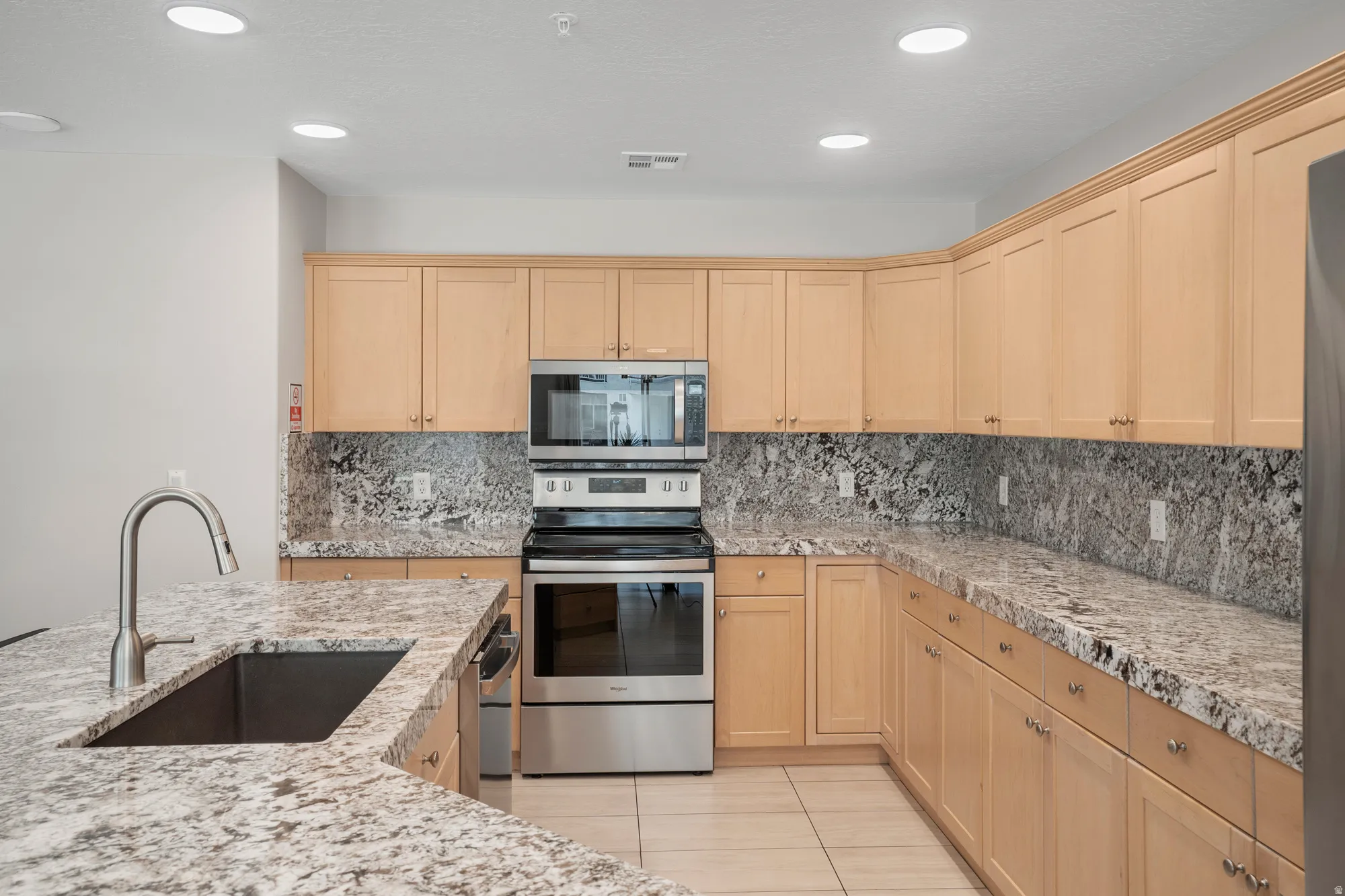 Spacious kitchen featuring light wood cabinetry, stone countertops, a stainless steel sink with a gooseneck faucet, and integrated stainless steel appliances
