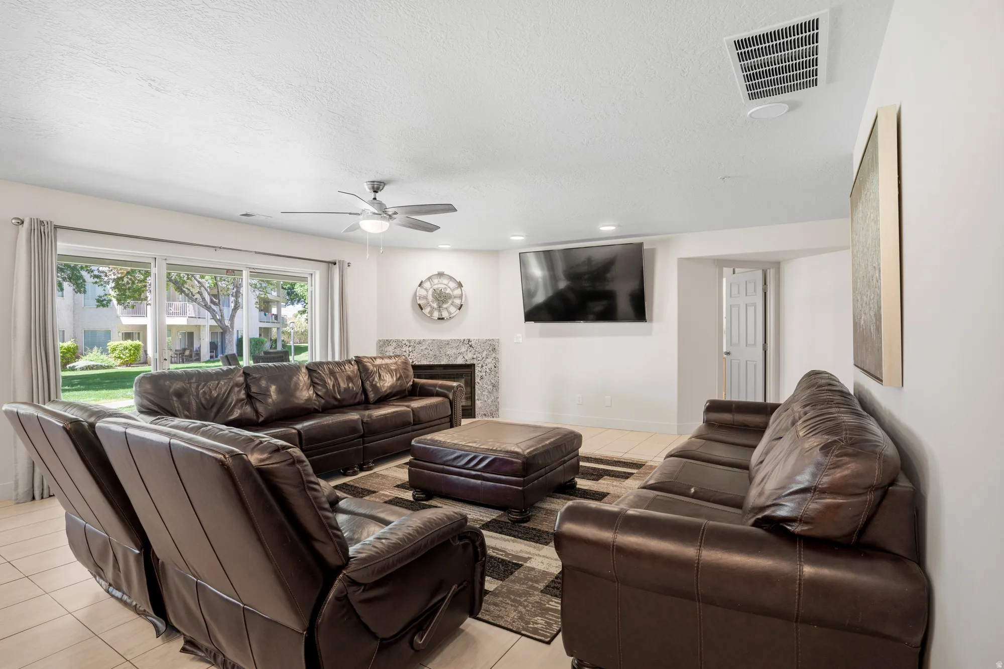 Spacious living area featuring light-toned tile flooring, a ceiling fan with integrated lighting, a fireplace with a stone surround, and large sliding glass doors providing access to the outdoors