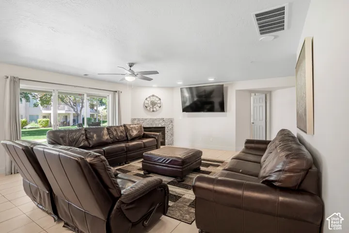 Spacious living area featuring light-toned tile flooring, a ceiling fan with integrated lighting, a fireplace with a stone surround, and large sliding glass doors providing access to the outdoors