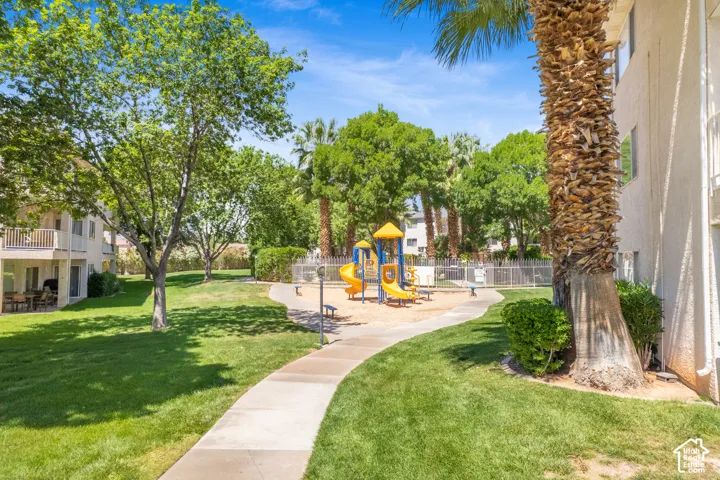 Landscaped outdoor area featuring a playground with slides and climbing structures, a concrete walkway, mature trees, green lawn, and a building exterior with balconies