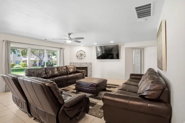 Spacious living area featuring light-toned tile flooring, a ceiling fan with integrated lighting, a fireplace with a stone surround, and large sliding glass doors providing access to the outdoors