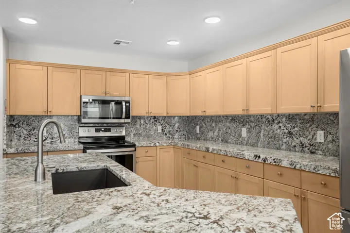 Kitchen featuring a stainless steel undermount sink with a gooseneck faucet, light wood cabinetry, granite countertops, a matching granite backsplash, and stainless steel appliances including a microwave and range