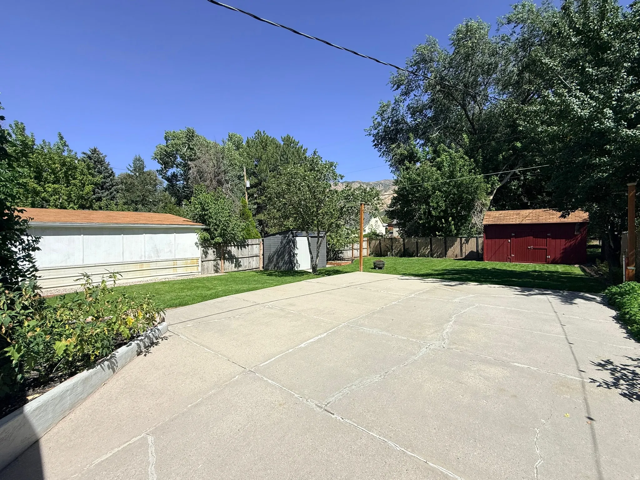 Spacious concrete pad, expansive grassy area, mature trees, wood fence, and a red storage shed with a shingled roof