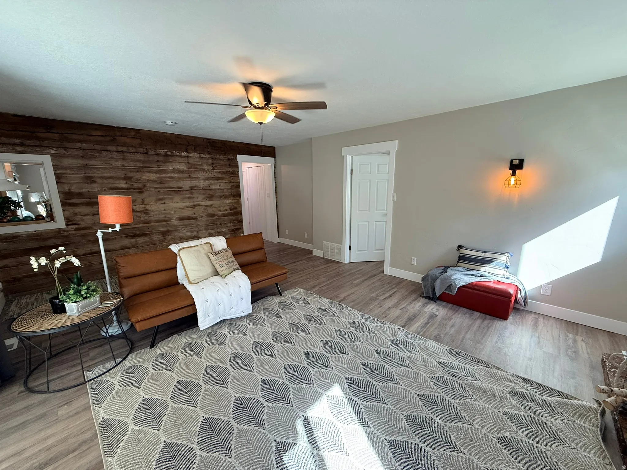 Wood-finish flooring, accent wall with horizontal wood paneling, ceiling fan with integrated lighting, and a wall-mounted sconce
