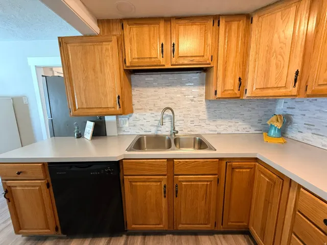 Wood-finish cabinetry with dark hardware, dual basin stainless steel sink, contemporary faucet, and mosaic tile backsplash