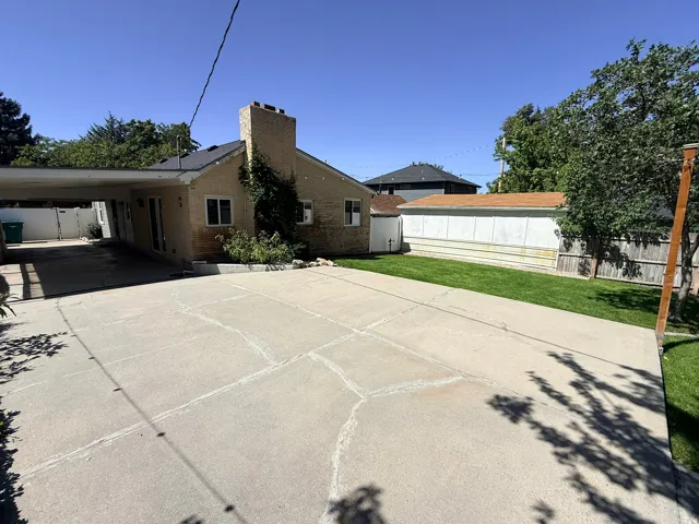 Brick exterior with a prominent chimney, attached carport, expansive concrete driveway, and a fenced perimeter